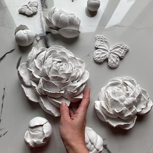 Op de afbeelding: Witte decoratieve wandkunstwerken met pioenrozen, bloemknoppen en vlindervormen. De bloemen- en vlinderontwerpen zijn gerangschikt op een marmeren oppervlak. De stukken zijn gemaakt van een effen, mat materiaal.