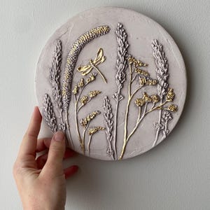 May include: A circular wall plaque in a light lavender shade featuring a raised, textured relief of botanical elements and a dragonfly. The plant stems and the dragonfly are highlighted with gold leaf accents, creating a contrast against the matte surface.