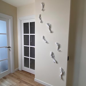 Pode incluir: Instalação de arte de parede com sete esculturas de peixes koi brancos dispostas verticalmente em uma parede creme. O corredor apresenta piso de madeira e uma porta com moldura branca.