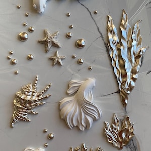 May include: A set of white and silver 3D wall decor featuring fish, starfish, seashells, and seaweed. The decor is perfect for a nautical themed room.