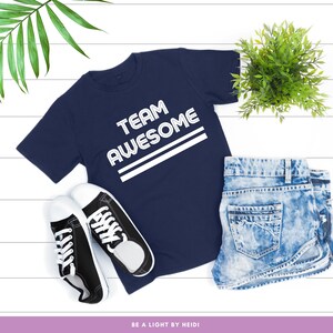 May include: A navy blue t-shirt with white text that reads "TEAM AWESOME" in a retro font. The shirt is laid flat on a white wooden surface with a pair of black canvas shoes and denim shorts.
