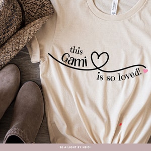 May include: A beige t-shirt with the text "this Gami is so loved!" in black with a pink heart. The shirt is on a wooden surface with a brown hat and brown boots.