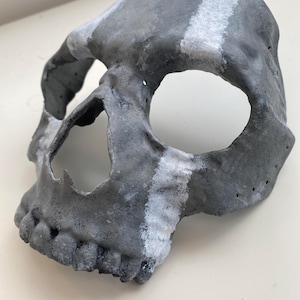 Puede incluir: Máscara de calavera gris con una franja blanca en la frente y la mejilla. La máscara tiene cuencas de ojos grandes y una mandíbula detallada. La máscara es probablemente para Halloween o disfraces.