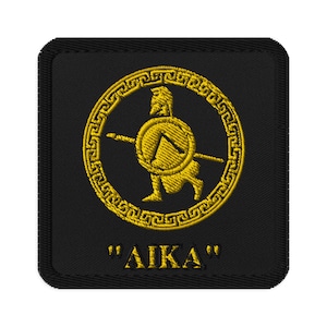 Puede incluir: Un parche bordado negro con un guerrero griego dorado sosteniendo una lanza y un escudo dentro de un círculo con un borde de clave griega. El texto "AIKA" está debajo del círculo.