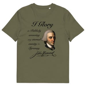 Peut inclure: T-shirt vert olive avec un portrait de John Hancock et la citation "I Glory in Publicly avowing my eternal enmity to Tyranny." Le design comprend la signature de Hancock en script noir.