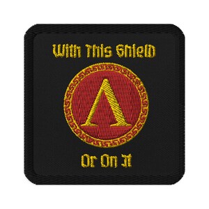 May include: Black embroidered patch with a gold circle and red shield with a yellow letter A inside. The text "With This Shield Or On It" is embroidered in yellow.