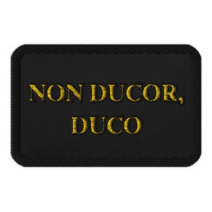 May include: Black rectangular patch with gold embroidered text that reads "NON DUCOR, DUCO". The patch has a stitched border and a textured appearance. The text is in a serif font.