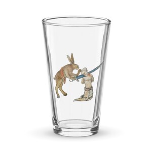 Könnte beinhalten: Ein klares Glas Pint-Glas mit einem gedruckten Bild eines Kaninchens, das ein Schwert hält und eine Person sticht. Das Kaninchen ist braun und die Person ist nackt mit einem roten Band um die Taille.