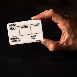 May include: A white film clapperboard with black text, labeled "Prod Roll", "Scene", "Take", "Camera Director", "Date", and "FPS". The clapperboard also has the text "OPE AK3" printed on the bottom.