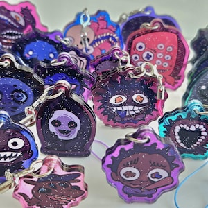 Puede incluir: Una colección de llaveros de acrílico con diversas ilustraciones de monstruos de dibujos animados y fondos galácticos brillantes en tonos morados, azules y rosas. Cada dije incluye una argolla de metal y un cordón de color.