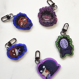 Può includere: Cinque portachiavi di personaggi dei cartoni animati con chiusure in metallo nero. I portachiavi presentano vari personaggi con diversi colori e design, tra cui un personaggio con un contorno viola e un personaggio con un contorno verde.