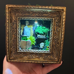 May include: A square, ornate gold-toned frame containing a miniature diorama. Inside, a green figure wearing a pointed hat holds an open book labelled "SPELLS". The scene includes a small skull, a black cauldron, green foliage, and a framed picture.