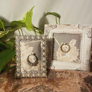 Op de afbeelding: Twee decoratieve ringhouders. Eén heeft een zilveren frame met imitatieparels en een ring. De andere heeft een wit verweerd frame met een gouden ring. Beide hebben bloemenstoffen achtergronden.