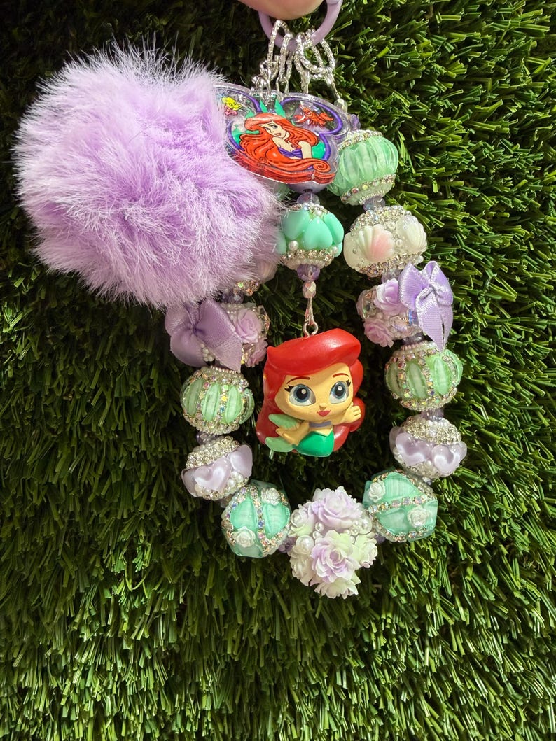 May include: A decorative keychain featuring a large lavender faux fur pom-pom, a central Ariel character charm, and a variety of pastel-colored beads including mint green, white, and purple accents, interspersed with small satin bows and rhinestones.