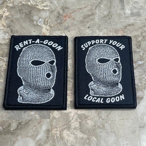 Può includere: Due toppe di tessuto nero con testo e immagini ricamate in bianco. La prima toppa dice "Rent-A-Goon" e presenta un'immagine ricamata in bianco di una persona che indossa una maschera da sci. La seconda toppa dice "Support Your Local Goon" e presenta un'immagine ricamata in bianco di una persona che indossa una maschera da sci.