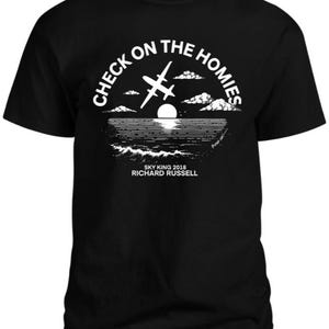 May include: Black t-shirt with a white graphic design of a plane flying over a sunset and ocean. The text "Check on the Homies" is in a circle around the plane. The text "Sky King 2018 Richard Russell" is below the graphic.
