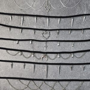 Może przedstawiać: Kolekcja czarnych chokerów ze srebrnymi wisiorkami i łańcuszkami. Wzory obejmują kolce, serca, krzyże i półksiężyce. Chokery są prezentowane na szarym tle, prezentując różnorodność biżuterii inspirowanej gotykiem.