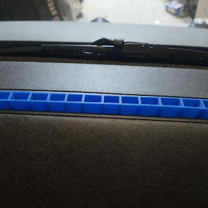 Può includere: Una copertura per bocchetta rettangolare blu brillante con più aperture è inserita in un cruscotto nero. Il tergicristallo è visibile sopra il cruscotto. La copertura della bocchetta è un accento vibrante contro l'interno scuro.