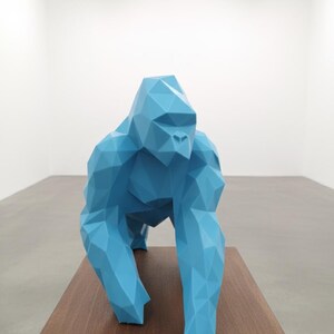 Könnte beinhalten: Eine geometrische, hellblaue Gorillaskulptur. Der Affe ist in einer quadrupeden Haltung dargestellt, mit einem facettierten Design. Sie wird auf einer dunkelbraunen Holzoberfläche vor weißem Hintergrund ausgestellt. Die Skulptur ist ein modernes Kunstwerk.