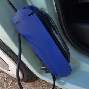 May include: A blue charging cable holder with a black charging cable. The holder is rectangular with curved edges and slots for the cable. The holder is mounted on a light blue car.