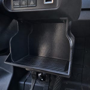 May include: A black plastic vehicle center console storage compartment with a ribbed base. Above the storage area, a control panel features three buttons with icons for windshield defrost, fan speed, and hazard lights, alongside a rectangular USB port.