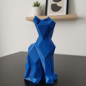 May include: A geometric, blue cat sculpture. The cat is in a seated position, with a faceted design. The sculpture is on a black surface, with a shelf, plant, and framed artwork in the background.