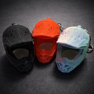 Peut inclure: Trois mini casques de moto en noir, orange et bleu-vert. Chaque casque a une visière et un porte-clés. Les casques sont imprimés en 3D avec des lignes de couche visibles, ce qui leur donne un aspect texturé. Le fond est une surface gris foncé.