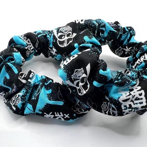 May include: Two black scrunchies with a repeating pattern of white and turquoise designs. The fabric is gathered, creating a textured appearance. The scrunchies are on a white surface.