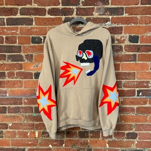 May include: A tan hoodie featuring a skull graphic with red glasses and flames. The sleeves have starburst designs in red, yellow, and blue. The hoodie is suspended on a grey hanger against a brick wall background.
