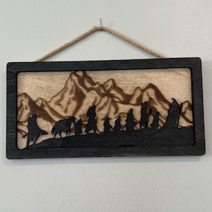 Può includere: Opera d'arte da parete in legno raffigurante un paesaggio montano con figure e animali. L'opera è incorniciata in nero e ha uno sfondo in legno naturale. Una corda è attaccata per appenderla.