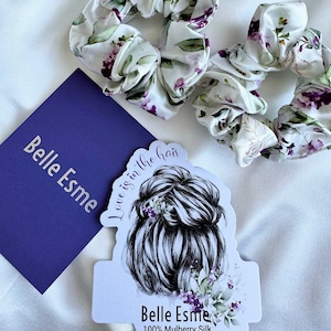 Puede incluir: Dos coleteros de seda blanca con un estampado floral de hojas moradas y verdes. Una tarjeta morada con el texto "Belle Esme" y una pegatina con un moño de mujer y el texto "Love is in the hair" también están en la imagen.