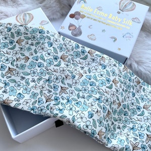Puede incluir: Una caja de regalo blanca con un tejido de estampado floral azul y blanco en su interior. La caja tiene una inscripción dorada que dice "Belle Esme Baby Silk" y "NEW!  100% CERTIFIED 22 MOMMES MULBERRY SILK THE GIFT OF LOVE FOR YOUR BABY"