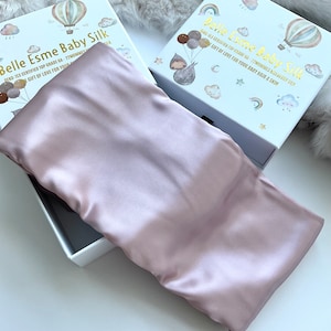Peut inclure: Un oreiller en soie rose pâle est présenté dans une boîte blanche avec des lettres et des illustrations dorées. La boîte indique "Belle Esme Baby Silk" et détaille la qualité du produit. L'oreiller en soie est un article doux et luxueux.