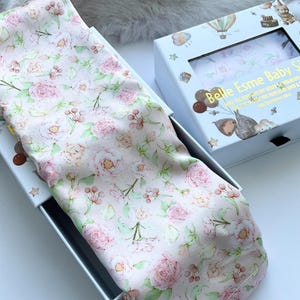 May include: A floral baby swaddle blanket with a pink rose pattern, partially visible inside a white box. The box has a clear window and the text "Belle Esme Baby Swaddle". The blanket is made of soft fabric.
