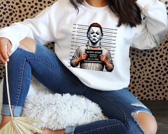 May include: White sweatshirt with a graphic of Michael Myers from the Halloween movie. The graphic shows Michael Myers in a white jumpsuit with a mugshot background. The text on the mugshot reads "MICHAEL MYERS, HADDONFIELD POLICE DEPT, OCT. 31, 1978".