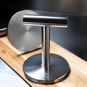 May include: A stainless steel burger press featuring a flat circular base, a vertical cylindrical handle, and a horizontal top bar. The base displays a circular logo with horizontal lines and the letters SM. The metal surface has a brushed finish.