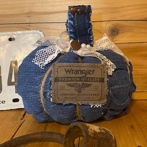 May include: A decorative pumpkin crafted from blue denim fabric, featuring a Wrangler brand patch on the front. The pumpkin is topped with a denim stem, white lace ribbon, and small pearl-like beads. The patch displays the text Wrangler Premium Quality Comfort Guaranteed.