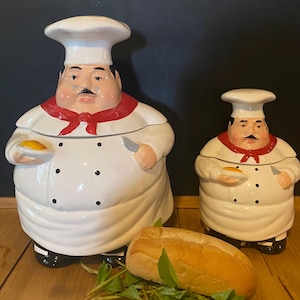 May include: Two ceramic chef-shaped canisters with white uniforms, red neckerchiefs, and black buttons. Each figure wears a tall white hat, has a black mustache, and holds a small plate with a yellow item and a silver knife in their hands.