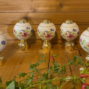 May include: Five spherical curtain rod finials made of white ceramic with a floral pattern featuring pink, white, and blue flowers. Each finial has a gold-toned metal base and a gold-toned cap at the top, with a gold-toned ring accenting the center.