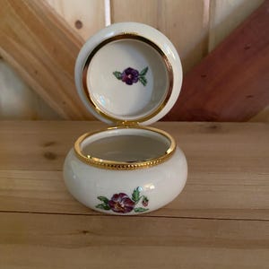May include: A white porcelain trinket box with a gold trim and a purple pansy flower design. The box is open and the inside is white.