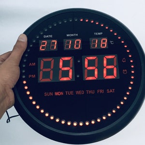 May include: A black round digital clock with a red LED display showing the time, date, month, and temperature. The clock has a ring of red LEDs around the edge.