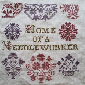 Op de afbeelding: Een witte stof met borduurwerk in kruissteek met verschillende bloemmotieven en de tekst "Home of a Needleworker".