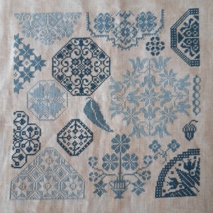 May include: A cross-stitch sampler with a variety of geometric and floral patterns in shades of blue. The design includes octagons, diamonds, and floral motifs, along with a bird and an acorn. The background is a light beige fabric.