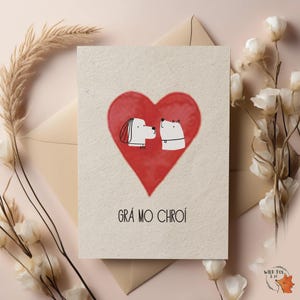 May include: A greeting card with a red heart and two cartoon dogs facing each other. The card has the text "Grá mo chroí" in black. The card is on a beige envelope, with flowers and decorative elements.