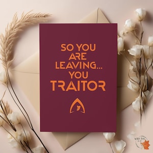 May include: A burgundy card with orange text: "SO YOU ARE LEAVING... YOU TRAITOR." The card features an orange emblem. It rests on a beige envelope, with white flowers and dried grass. A humorous farewell card.