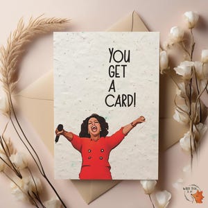 May include: Greeting card with the text "YOU GET A CARD!" and an illustration of a woman in a red outfit holding a microphone with her arms outstretched. The card has a textured, off-white background.