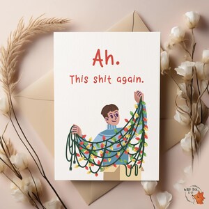 May include: A greeting card with the text "Ah. This shit again." features an illustration of a person holding Christmas lights. The card has a beige envelope and is surrounded by flowers and foliage.