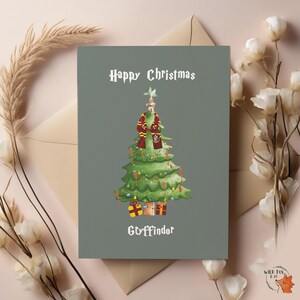 Puede incluir: Una tarjeta de Navidad con un fondo verde. Presenta una ilustración en acuarela de un árbol de Navidad adornado con luces y regalos. Una bufanda y una pequeña figura decoran el árbol. La tarjeta dice "Happy Christmas" y "Gryffindor".