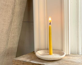 May include: A lit yellow candle in a small, off-white, speckled dish. The candle is centered in the dish and is burning with a bright flame. The candle and dish are on a wooden surface, with a white window frame in the background.