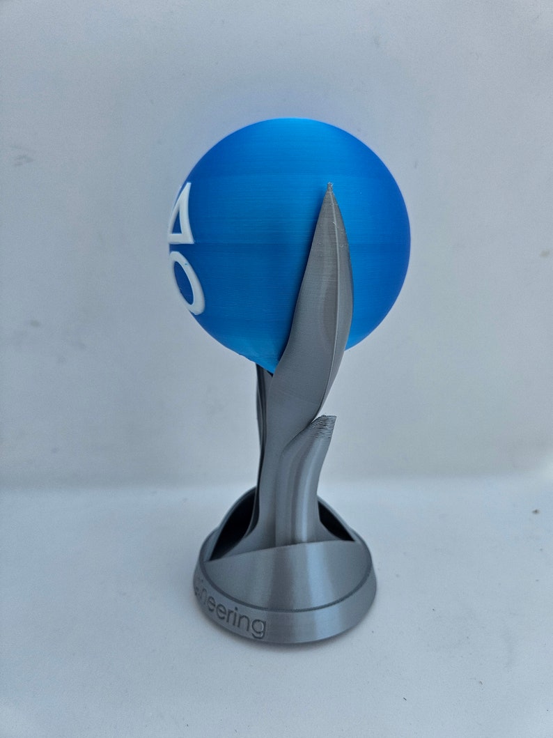 May include: A 3D printed trophy with a blue sphere on top. The sphere has a white logo with the letters "AO" on it. The base of the trophy is gray and has the word "Engineering" inscribed on it.